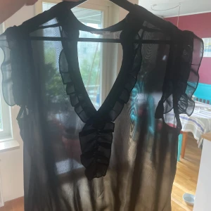 Svart transparent blus med volangdetaljer - Säljer en elegant svart blus i transparent material. Den har fina volangdetaljer längs med halsringningen och ärmlösa axlar. Perfekt för en festlig kväll eller en stilfull dag på stan.