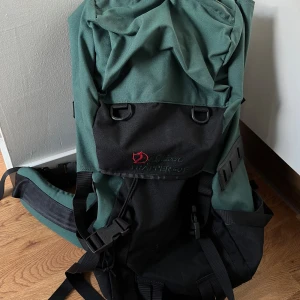 Fjällräven Trapper 50F ryggsäck - Fjällräven Trapper 50F ryggsäck i grönt och svart. Ryggsäcken har en stor huvudficka med dragskoöppning och en mindre ficka på framsidan. Den har även ett justerbart bröstband och ett höftbälte. Ryggsäcken är tillverkad av slitstarkt material och är 
