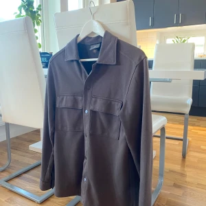 Brun overshirt från H&M - Säljer en snygg brun overshirt från H&M i storlek S. Perfekt för höst och vår med sin stilrena design och långa ärmar. Skjortan har knappar framtill och två bröstfickor som ger den ett intressantare utseende. Passar både till vardags och till fest! Bara att kontakta mig vid eventuella frågor!