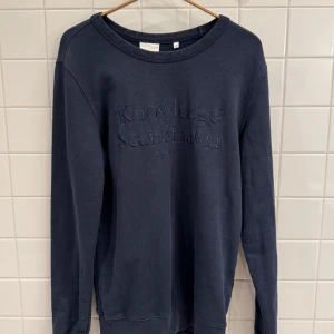 Mörkblå tröja från Knowledge Cotton Apparel - Säljer en snygg mörkblå tröja från Knowledge Cotton Apparel. Den är i bomull och har en stilren design med texten 'Knowledge Scandinavia' på framsidan. Perfekt för både vardag och chill. Tröjan har långa ärmar och en rund halsringning.