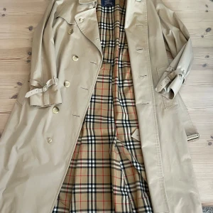 Beige trenchcoat från Burberrys - Säljer en klassisk beige trenchcoat från Burberrys. Den har ett ikoniskt rutigt mönster på insidan och är utrustad med knappar och ett bälte vid ärmsluten. Perfekt för höst och vår! Storlek M 