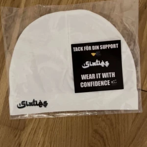 Vit mössa med tryck - Säljer en snygg vit mössa med ett coolt tryck framtill. Perfekt för att hålla sig varm och stilren under kallare dagar. Mössan är ny och kommer i originalförpackning. Bär den med självförtroende!