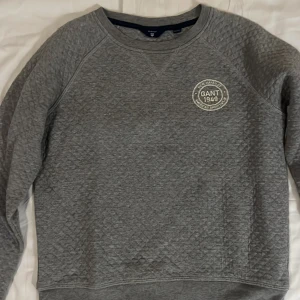 Grå quiltad tröja från GANT - Säljer en stilren grå quiltad tröja från GANT med rund halsringning och ett broderat emblem på bröstet. Perfekt för kyligare dagar med sin mjuka och bekväma passform. Passar barn i åldern 9-10 år.