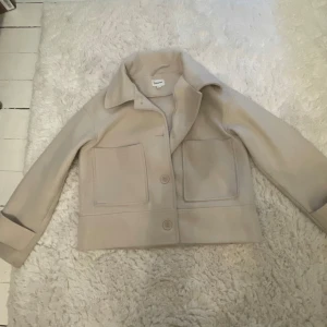 Beige jacka från Kappahl - Snygg beige jacka från Kappahl med stora fickor och knappar framtill. Jackan har en klassisk krage och är perfekt för höst och vår. Den är tillverkad i ett mjukt material som ger en stilren look. Köpt för 900kr