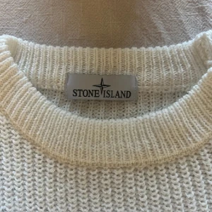 Vit stickad tröja från Stone Island - Säljer en stilren vit stickad tröja från Stone Island. Tröjan har en klassisk rund halsringning och det ikoniska Stone Island-märket på ärmen. Perfekt för att hålla sig varm och snygg under kyligare dagar.