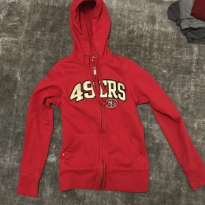 Röd hoodie från Antigua - Säljer en röd hoodie från Antigua med 49ers-tryck på framsidan. Den har en dragkedja och en luva. 