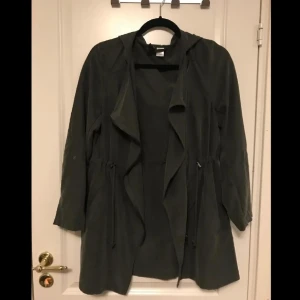 Grön och brun trenchcoat med huva - En mörk grön och en brun trenchcoat med huva och dragsko i midjan. Jackan har långa ärmar och en avslappnad passform. Perfekt för höst och vår. 80kr styck eller båda för 120kr