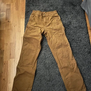 Bruna arbetsbyxor från Carhartt - Säljer ett par robusta bruna arbetsbyxor från Carhartt. Byxorna har flera praktiska fickor och en klassisk design med hög midja. Perfekta för utomhusarbete eller en avslappnad stil. Sitter mer som 28 eller x 32.