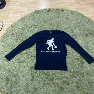 Mörkblå långärmad tröja från Name It - Säljer en mörkblå långärmad tröja från Name It med ett tryck av en basketspelare och texten 'Athletic Champion' på framsidan. Perfekt för sportiga tillfällen eller som en cool vardagströja. Passar åldrarna 11-12 år.