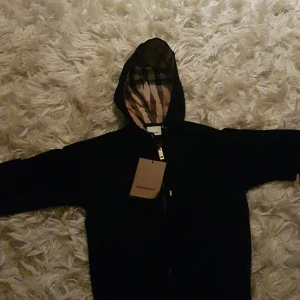 Svart jacka från Burberry - Säljer en stilren svart jacka från Burberry med klassiskt rutigt mönster i luvan. Jackan har dragkedja framtill och en elegant design som passar perfekt för olika tillfällen.