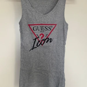 Grå ribbad linnetopp från Guess - Snygg grå ribbad linnetopp från Guess med det ikoniska röda och svarta logotrycket framtill. Perfekt för en avslappnad stil med en touch av elegans.