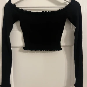 Svart offshoulder topp - Snygg svart offshoulder topp med långa ärmar och ribbad design. Toppen är croppad och har en elastisk kant vid halsen för en bekväm passform. Perfekt för en stilren look.