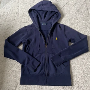 Mörkblå hoodie från Ralph Lauren - Säljer en snygg mörkblå hoodie från Ralph Lauren med dragkedja och det klassiska logotypbroderiet på bröstet. Perfekt för en avslappnad stil. Den har långa ärmar och en praktisk huva med dragsko. Ska kunna passa xs med!