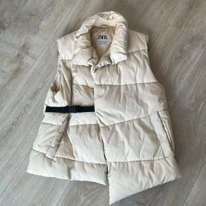 Beige dunväst från Zara - Säljer en snygg beige dunväst från Zara med ett praktiskt bälte i midjan. Jag är 160 cm , Västen har en hög krage och är perfekt för kyligare dagar. Den är quiltad och ger en stilren look.