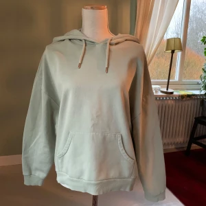Pastellgrön hoodie från Lindex - Säljer en pastellgrön hoodie från Lindex i storlek L. Den har en klassisk design med en stor ficka framtill och justerbar huva med snören. Perfekt för en avslappnad stil. Tröjan är i fint skick utan noppror och fläckar