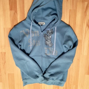 Blå hoodie från Chasin' Butterflies - Snygg blå hoodie från vonstudio med dragkedja och glittrig text och motiv på framsidan. Den har en justerbar huva med snören och ribbade muddar vid ärmslut och nederkant. Perfekt för en avslappnad stil.