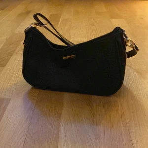 Svart axelväska med gulddetaljer - Snygg svart axelväska med gulddetaljer och dragkedja. Perfekt för att bära dina nödvändigheter med stil. Väskan har en elegant design och en justerbar axelrem.