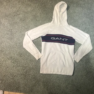 Vit hoodie från GANT - Säljer en stilren vit hoodie från GANT med en blå och röd rand över bröstet och GANT-loggan. Tröjan har en klassisk känguruficka och en bekväm huva. Perfekt för en avslappnad stil.
