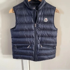Mörkblå dunväst från Moncler - Moncler gui väst strl 14 (modellen är 167cm).Använd fåtal gånger utan defekter .Västen är perfekt till kyligare dagar. Den är stilren och lätt att matcha med olika outfits. Ny pris: 5000kr. Mitt pris: 2500. Bara att höra av sig vid minsta fundering. 