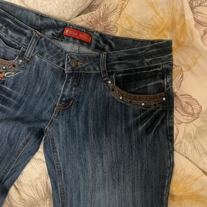 Blå low waisted bootcut jeans - Snygga blå jeans med unika broderade detaljer och nitar vid fickorna. De är bootcut och low waisted. Perfekta för dig som vill ha något extra i garderoben! Säljer då de är för stora på mig skulle sjukt gärna haft kvar dem annars💕