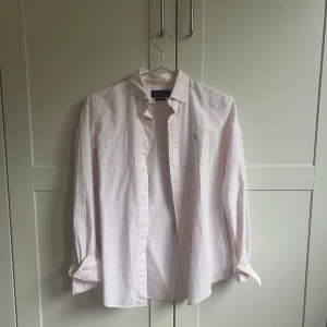 Rosa och vit randig skjorta från Ralph Lauren - Säljer en stilren rosa och vit randig skjorta från Ralph Lauren i ”slimfit”, storlek XS. Skjortan har långa ärmar och klassiska knappar framtill. Säljer för 300kr + frakt 💫🫶🏻💕