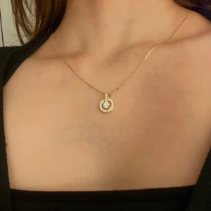 Guldhalsband med glittrig hängsmycke - Elegant guldhalsband med ett cirkulärt hängsmycke dekorerat med gnistrande stenar. Perfekt för att ge en touch av glamour till din outfit. Kedjan är tunn och smidig med en säker låsning. Från ”snö”.