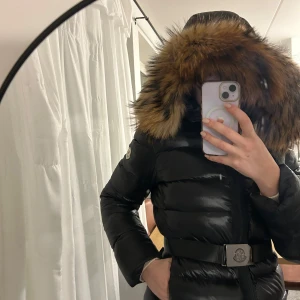 Svart dunjacka med pälsdetalj från Moncler - Snygg svart dunjacka från Moncler med pälsdetalj på huvan och ett bälte i midjan. Jackan har en dragkedja framtill och långa ärmar. Perfekt för kalla dagar när du vill hålla stilen. Kan användas med eller utan bälte. Storleken är en XS-S beroende på hur man vill ha den. Jag upplever den som liten så skulle personligen säga XS. Men storlek 3 