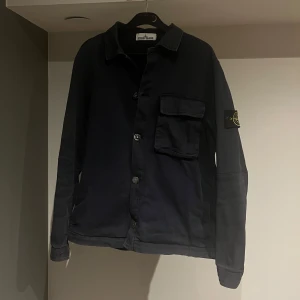 Svart overshirt från Stone Island - Riktigt fet mörkblå overshirt från Stone Island med klassisk krage och knappstängning. Jackan har en bröstficka och den ikoniska logotypen på ärmen. Perfekt till våren eller sommaren med en t-Shirt under! Storlek S. Jag själv är 180 cm och väger 72kg, passar perfekt. Jackan är använd, men i relativt bra skick. Bara att skriva om du har eventuella frågor!😉