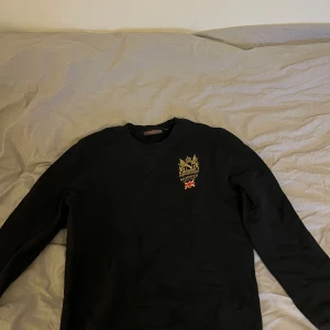 Svart tröja från Morris - Snygg svart tröja från Morris med ett broderat emblem och brittisk flagga på bröstet. Perfekt för en stilren look. Tröjan har långa ärmar och rund halsringning.