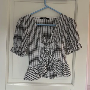 Rutig blus från Bik Bok - Säljer en söt rutig blus från Bik Bok i svart och vitt. Blusen har en v-ringad design med knytning framtill och volangdetaljer vid ärmslut och nederkant. Perfekt för en stilren look!