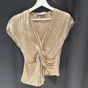 Beige plisserad blus från Zara - Elegant beige blus från Zara med plisserad design och omlott framtill. Blusen har korta ärmar och en v-ringad halslinje, vilket ger en stilren och modern look.