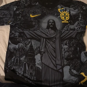 Svart Brasilien fotbollströja med tryck - Snygg svart fotbollströja från Nike med Brasilien-tema. Tröjan har ett unikt tryck med Kristusstatyn och andra detaljer i grått och gult. Den har korta ärmar och Brasilien-emblemet på bröstet. Perfekt för fotbollsfans som vill sticka ut!