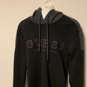 Svart och grå hoodie från Guess - Snygg svart hoodie från Guess med grå detaljer och texten 'Guess Los Angeles' på framsidan. Den har en justerbar huva med dragsko och långa ärmar. Perfekt för en avslappnad stil. Storlek S.