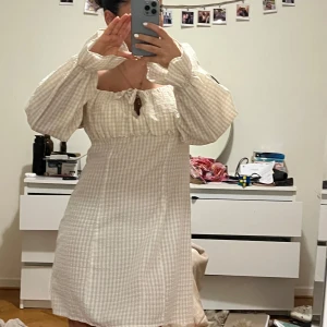Beige rutig klänning - Charmig beige rutig klänning med långa puffärmar och en söt knytning framtill. Perfekt för en avslappnad och stilren look. Den har en kort längd och använt 2 gånger.