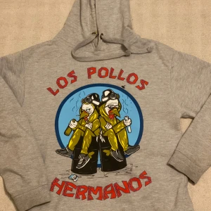 Grå hoodie med Los Pollos Hermanos tryck - Säljer en grå hoodie från Just Hoods med ett coolt Los Pollos Hermanos tryck på framsidan. Tröjan har en klassisk huva med dragsko och långa ärmar. Perfekt för fans av Breaking Bad!