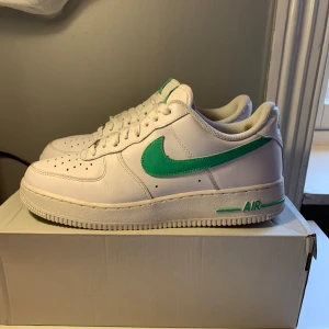 Nike Air Force med grön swoosh - Snygga Nike Air Force sneakers i vitt med en grön swoosh och gröna detaljer. Skorna har klassisk snörning och perforeringar på tån för ventilation. Perfekta för en stilren look med en färgklick.