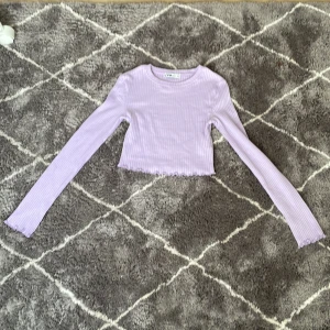 Ljuslila ribbad topp från New Yorker - Säljer en söt ljuslila ribbad topp från New Yorker. Den är croppad med långa ärmar och har en fin vågig kant vid ärmslut och nederkant. Perfekt för en stilren look! 💜