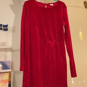 Röd klänning med glittriga detaljer - Snygg röd klänning i sammet med glittriga prickar. Den har långa ärmar och en bekväm passform som passar perfekt för festliga tillfällen. Klänningen har en enkel design med en lätt markerad midja.