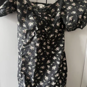 Svart blommig klänning - Charmig svart klänning med små blommor i rosa och grönt. Klänningen har korta ärmar och en rynkad detalj framtill som ger en fin form. Perfekt för en somrig look!