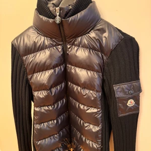 Svart pufferjacka/ cardigan  från Moncler - Snygg svart pufferjacka från Moncler med stickade ärmar och hög krage. Jackan har en dragkedja framtill och en ficka med knappdetalj på ärmen. Perfekt som vår/ höst jacka. Plagget är aldrig använd och orginal box medföljer. 