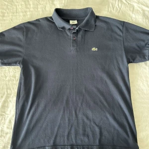Lacoste Polo - Säljer en klassisk mörkblå pikétröja från Lacoste med den ikoniska krokodilloggan på bröstet. Tröjan har korta ärmar och knappar vid kragen. Perfekt för en stilren och avslappnad look.