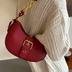Röd axelväska med guldkedja - Snygg röd axelväska i skinn med en elegant guldkedja och justerbart bälte. Perfekt för att ge din outfit en färgklick. Väskan har en unik form och stilrena detaljer.