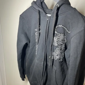 Svart hoodie med tigerdesign -  svart hoodie från Von studio  med en glittrig tigerdesign på framsidan. Tröjan har dragkedja och huva. Storlek M. Säljs för billigt pris. Vid fundering kontakta gärna oss. 
