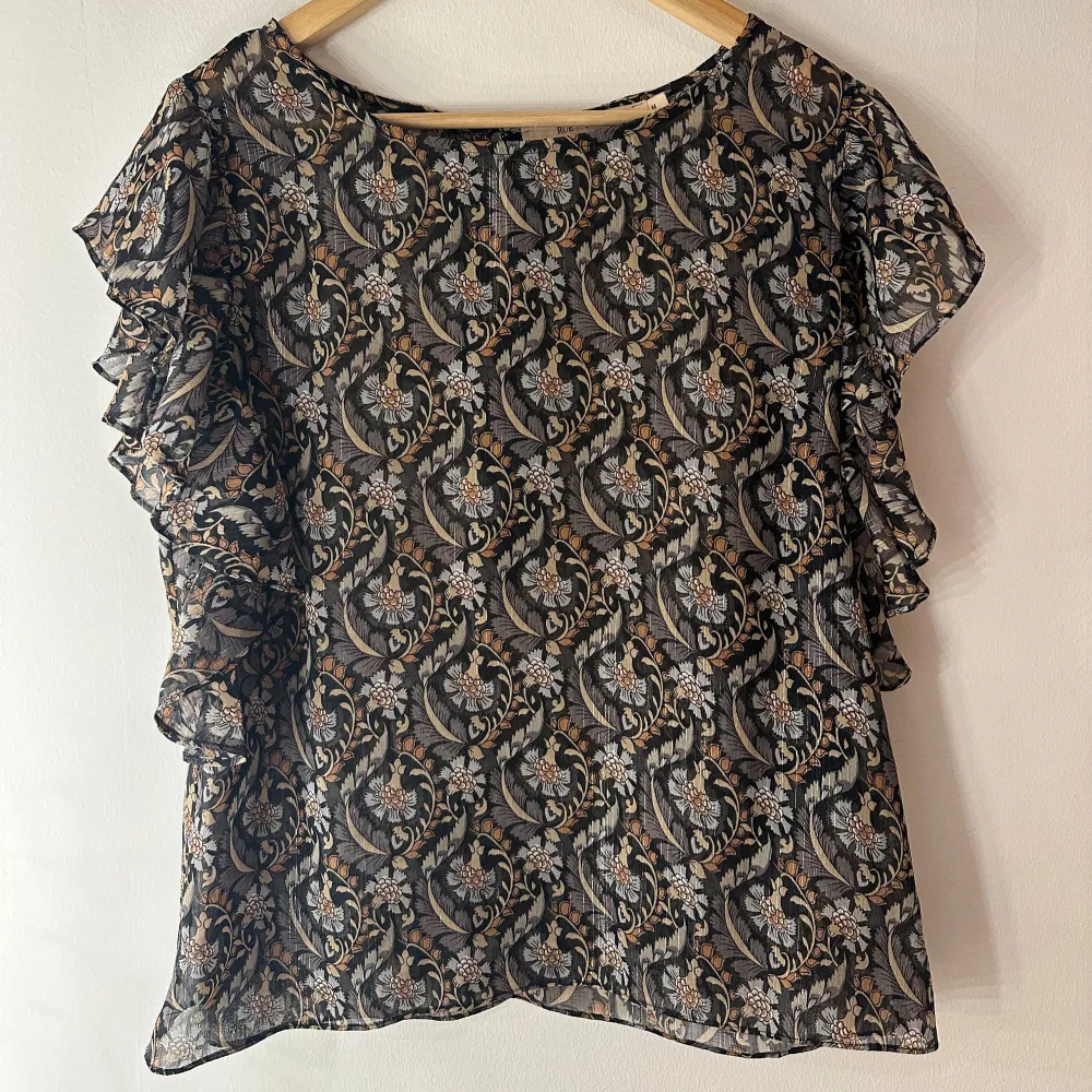 Snygg och elegant blus från Rue de Femme med ett vackert blommigt mönster i svart, beige och grå toner. Blusen har en avslappnad passform med volangdetaljer vid ärmarna som ger den en romantisk touch. Rund halsringning och knappar i ryggen. Perfekt för både vardag och fest. Något genomskinlig så kan behövas ett linne under.. Puserot.