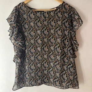 Blus  - Snygg och elegant blus från Rue de Femme med ett vackert blommigt mönster i svart, beige och grå toner. Blusen har en avslappnad passform med volangdetaljer vid ärmarna som ger den en romantisk touch. Rund halsringning och knappar i ryggen. Perfekt för både vardag och fest. Något genomskinlig så kan behövas ett linne under.