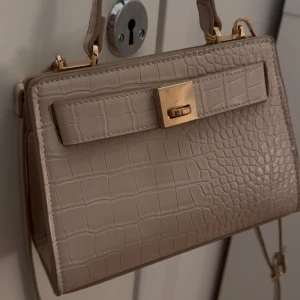 Beige handväska med krokodilmönster - Snygg beige handväska med krokodilmönster och gulddetaljer. Väskan har en elegant design med ett dekorativt spänne framtill och en praktisk axelrem. Perfekt för att ge din outfit en stilfull touch😍😍