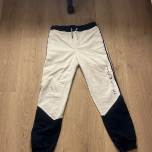 Vit och marinblå mjukisbyxor från Tommy Hilfiger - Säljer ett par snygga mjukisbyxor från Tommy Hilfiger i vit och marinblå färg. Byxorna har en elastisk midja med snörning och en cool randig detalj längs sidorna. Perfekta för en avslappnad stil. Står att de passar mellan åldrar 16-18 men tycker själv att de sitter som S