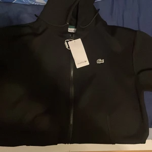 Svart hoodie från Lacoste - Snygg svart hoodie från Lacoste med dragkedja och den ikoniska krokodilloggan på bröstet. Perfekt för en stilren och avslappnad look. Har haft den ett bra tag men aldrig använt den och den har bara stått k garderoben den är storlek XL men funkar på de som använder L det kan vittna.