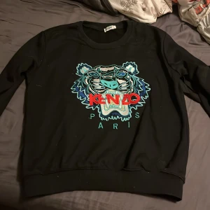 Svart sweatshirt från Kenzo - Snygg svart sweatshirt från Kenzo med det ikoniska tigerbroderiet i blått och rött på framsidan. Perfekt för en stilren och trendig look. Rund hals och långa ärmar. Storlek S men är liten i storleken