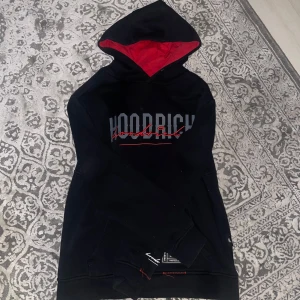 Svart hoodie från Hoodrich - Snygg svart hoodie från Hoodrich med röd text och detaljer. Den har en röd insida i huvan och en stor ficka framtill. Perfekt för en casual look.